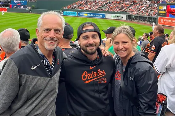 Dr. Yalich and two people smiling at a baseball game, one wearing an Orioles hoodie. They're surrounded by other fans, with the field and stadium visible in the background.