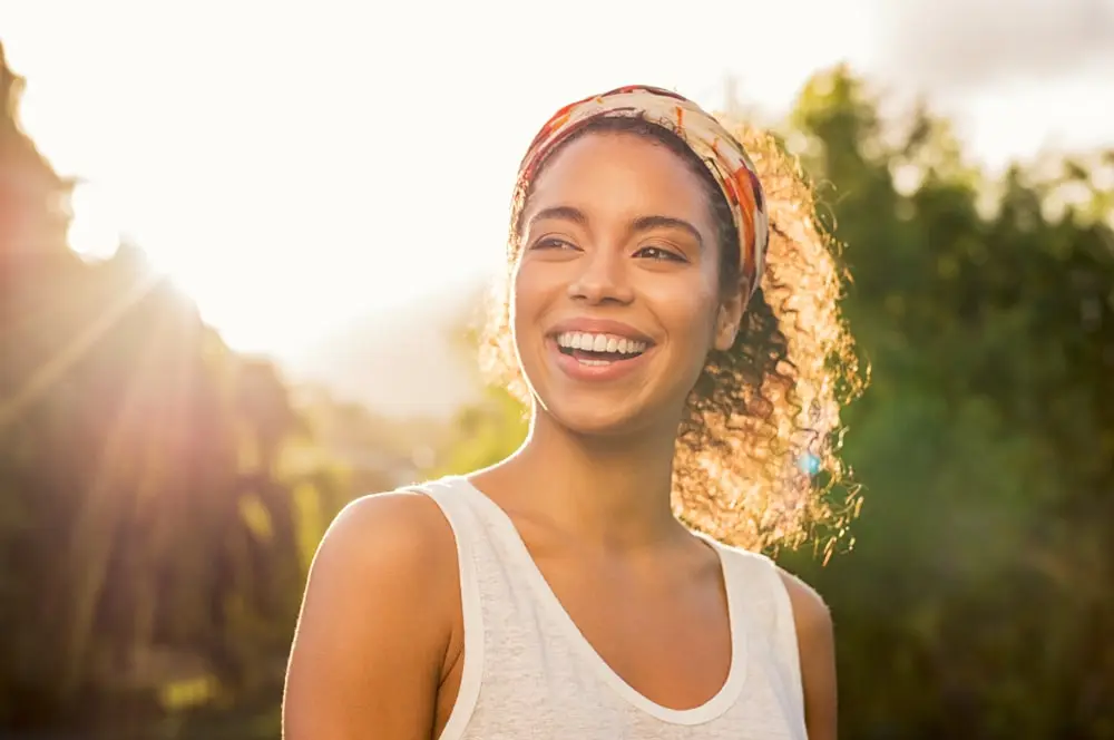 A woman with curly hair and a headband smiles warmly while standing outdoors in the sunlight. She is wearing a sleeveless top, and the background features greenery and soft sunlight filtering through trees.