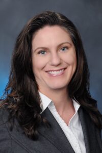 A woman with long brown hair wearing a white blouse and a gray blazer smiles at the camera against a dark, softly blurred background.