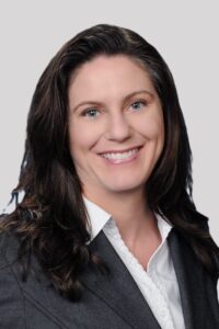 A woman with long, wavy dark hair wearing a gray blazer and white shirt smiles in front of a plain light gray background.