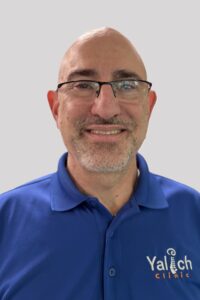 A smiling man with glasses, a bald head, and a trimmed beard wearing a blue polo shirt embroidered with Yalech Clinic stands against a light gray background.