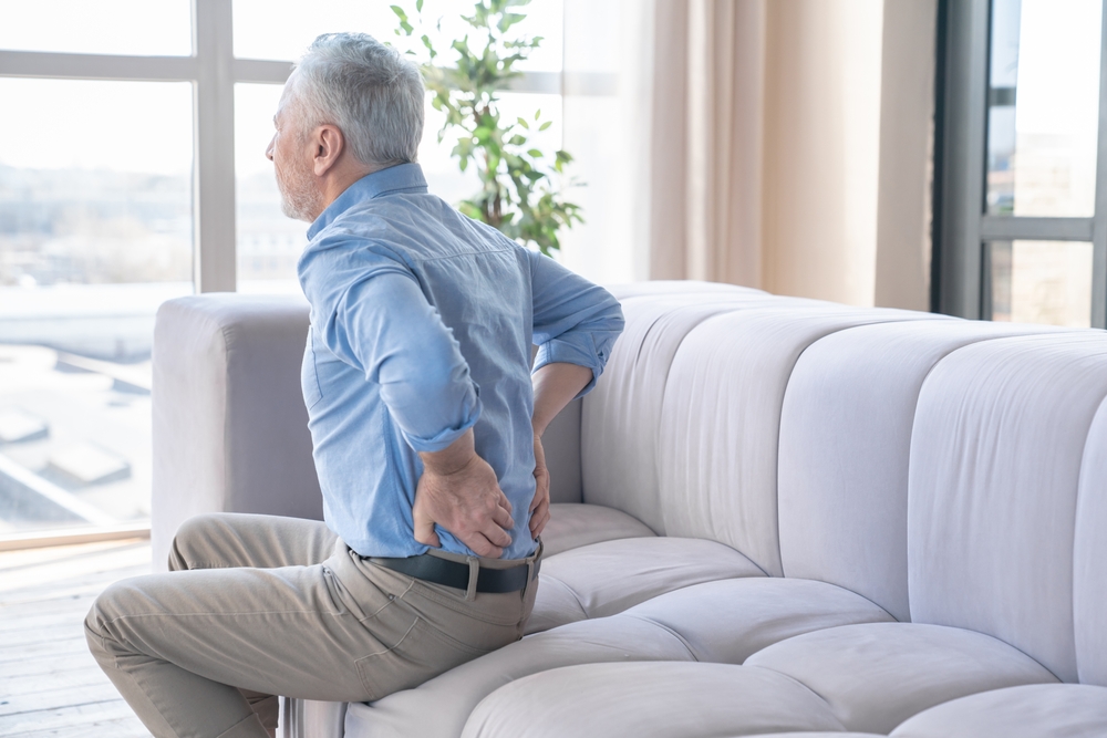 An older man sits on a sofa holding his lower back with both hands, appearing to be in pain or discomfort, in a bright living room with large windows.