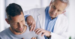 A doctor examines a mans shoulder, gently pressing as the man winces in pain. The doctor wears a white coat and blue shirt, while the man wears a gray sweater. The setting appears to be a medical office.