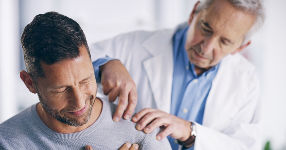 A doctor examines a mans shoulder, gently pressing as the man winces in pain. The doctor wears a white coat and blue shirt, while the man wears a gray sweater. The setting appears to be a medical office.