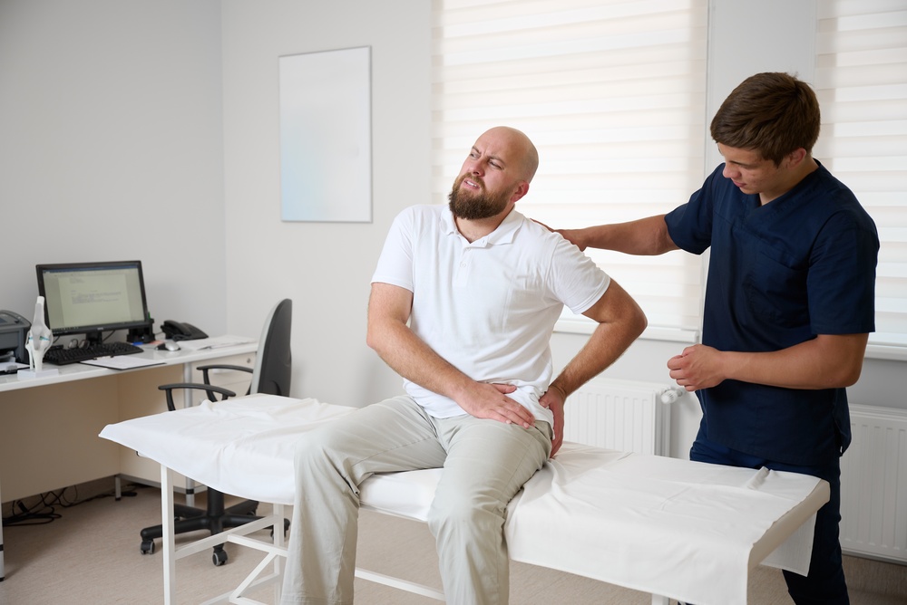 A man sits on an exam table holding his lower back in pain while a healthcare professional examines him in a medical office.