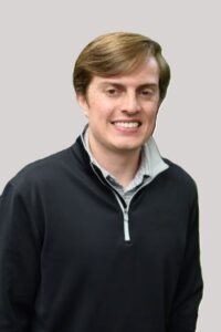 A young man with light brown hair wearing a black pullover with a gray zipper and a light-colored collared shirt underneath, smiling at the camera against a plain light background.