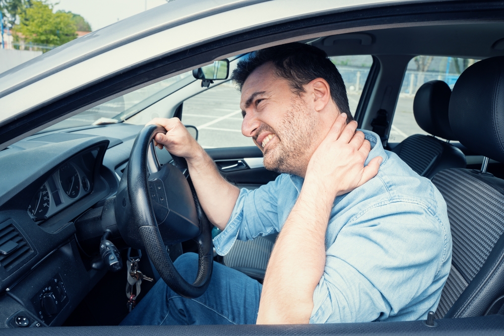 A man sitting in the driver’s seat of a car holds his neck in pain with one hand while gripping the steering wheel with the other, appearing to suffer from whiplash or neck discomfort.