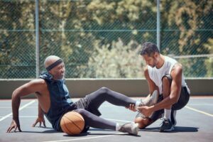 A man sits on an outdoor basketball court holding his ankle in pain while another man kneels beside him, checking his injury. A basketball rests near the injured man.